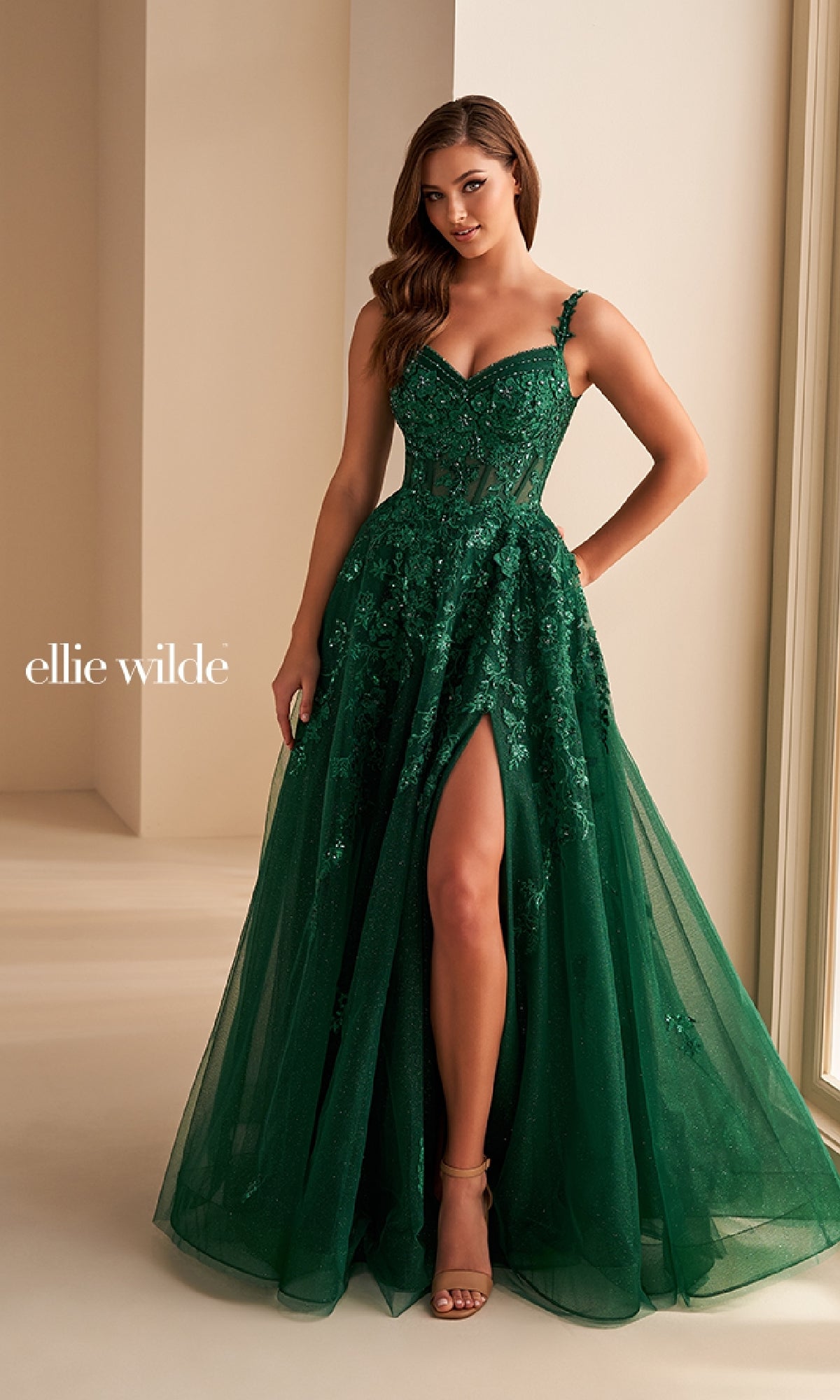 A woman wears the Ellie Wilde EW36204, a sleeveless emerald green long prom dress with floral lace appliques and a thigh-high slit, standing indoors near large windows.
